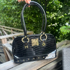 Svart handväska från Pompoos - Snygg svart handväska från Pompoos med croco-mönstrad yta i glansigt skinn. Väskan har guldiga detaljer, stora ringar och bokstäverna H och G framtill. Insidan är fodrad i starkt rosa tyg med flera fack och dragkedja. Perfekt för dig som gillar statement-accessoarer. Den är som stortsätt ny och inga tydliga märken. 