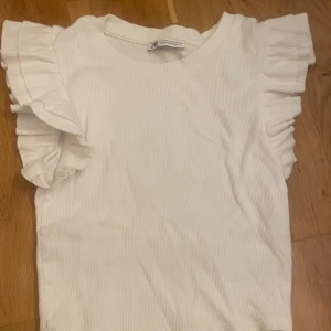 Vit volangtopp - Supersöt vit topp från Zara i ribbad bomull med volangdetaljer på axlarna. Modellen är ärmlös och har en rund halsringning. I storlek S men passar mer som en XS. Knappt använd och i superbra skick!!😍