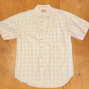 Beige rutig skjorta Pur et Simple - Kortärmad skjorta från Pur et Simple i ljusbeige med diskret rutigt mönster. Skjortan har klassisk krage, knappar framtill och en bröstficka. Tillverkad i en skön blandning av bomull och linne, perfekt för varmare dagar.