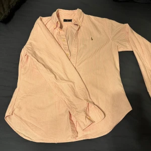 Rosa och vit randig skjorta Ralph Lauren - Säljer en klassisk rosa och vit randig skjorta från Ralph Lauren i 100% bomull. Skjortan har lång ärm, knappar framtill och den ikoniska broderade loggan på bröstet. Perfekt för dig som gillar preppy stil och vill ha något snyggt och tidlöst i garderoben.