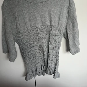 Grå smockad topp från H&M - Säljer en grå topp från H&M i storlek S. Toppen har trekvartsärm och ett ribbat, smockat parti över magen som ger en cool och unik look. Materialet är mjukt och stretchigt, perfekt för dig som gillar bekväma och stilrena plagg.
