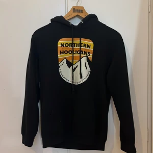 Svart hoodie Northern Hooligans XS - Svart hoodie från Northern Hooligans i storlek XS. Snyggt tryck framtill med berg och text i gult, vitt och svart. Klassisk huva med snörning och långa ärmar. Perfekt för chill dagar och streetwear-stil.