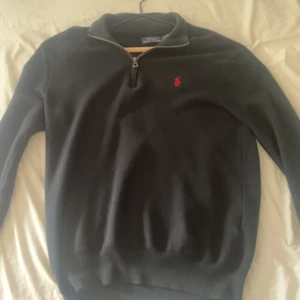 Svart half zip tröja från Ralph Lauren - Svart långärmad tröja från Ralph Lauren med half zip och klassisk röd logga broderad på bröstet. Tröjan har en stilren krage och är gjord i mjukt bomullsmaterial. Perfekt för dig som gillar en clean och tidlös look. Tröjan är perfekt nu till hösten och är i bra skick 