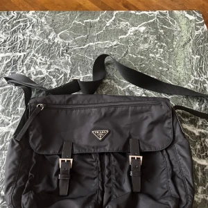 Prada Messenger bag - Snygg svart axelväska från Prada i slitstark nylon med justerbar rem, två framfickor med spännen och dragkedja på locket. Klassisk trekantslogga framtill och innerfoder med Pradas mönster. Perfekt för dig som vill ha en stilren och praktisk väska. Läderna flärparna är utbytta samt att tyget har lossnat inuti ena facket men är enkelt att fixa för den som orkar. 