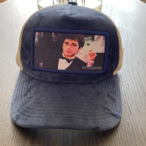 Blå och beige keps från Maggiore - Snygg keps från Maggiore med blå front i mjukt tyg och beige mesh baktill. Framsidan har en stor patch med foto och texten 'MAGGIORE'. Justerbar rem bak i svart. Perfekt för dig som vill sticka ut med en unik accessoar.