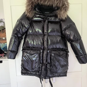 Svart glansig dunjacka Peak Performance - Nyskick! Aldrig använd! Snygg svart glansig dunjacka från Peak Performance med stor fluffig fuskpäls på huvan. Jackan har dragkedja, knappar, stora fickor och snörning i midjan. Perfekt för kalla dagar när du vill vara både varm och stilren. Storlek xs men passar även S skulle även tro M. Nypris: 6549:- 