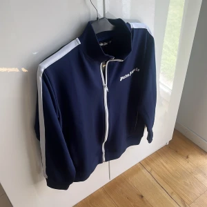 Palm Angels tröja - Snygg marinblå vindjacka från Palm Angels med vita ränder längs ärmarna och vit dragkedja. Jackan har hög krage, broderad logga på bröstet och sportig vibe. Perfekt för dig som gillar streetwear och vill sticka ut med stilrena detaljer. Mvh ItsForfits🌟