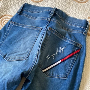 Tommy Hilfiger blå jeans med dragkedjor - Snygga blå jeans från Tommy Hilfiger med broderad logga och röd, vit och marinblå detalj på bakfickan. Jeansen har dragkedjor vid bensluten. Perfekta för dig som vill ha en cool och unik denimlook.