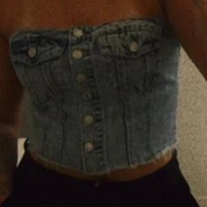 Jeans topp - Säljer en blå jeans bandeau tubtopp med snygga silverfärgade knappar framtill och två fejkade bröstfickor. Toppen har rå fransad nederkant och smockad baksida för extra stretch. Perfekt för dig som vill ha en cool och trendig denimlook.