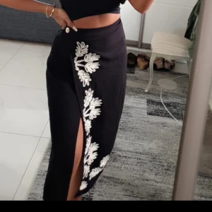 Svart omlottkjol med broderi - Svart lång omlottkjol med vit broderad blommönster längs kanten och en snygg slits framtill. Kjolen har en knapp i midjan och är figurnära med en elegant siluett. Perfekt för dig som vill sticka ut med coola detaljer.