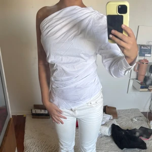 Vit oneshoulder topp med knappar - Säljer en vit oneshoulder topp med lång ärm på ena sidan och snygga knappar längs ryggen. Toppen har en tight passform. Ger en ”classy” och stilren look, går att använda både till vardags och som festtopp. 