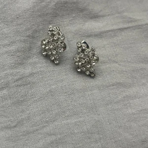 Glittriga silvriga örhängen - Säljer ett par snygga örhängen i silverfärg med glittrande stenar. De har en cool, snirklig form och är täckta av små kristaller som verkligen fångar ljuset. Perfekta för dig som vill sticka ut och addera lite extra bling till din look.