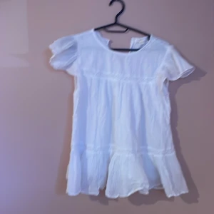 Vit blus från Gina Tricot - Supersöt vit blus från Gina Tricot i storlek 158/164. Blusen har korta puffärmar, volang nedtill och en mjuk, luftig känsla. Perfekt för varma dagar med sin flowiga passform och tunna bomullsmaterial. Rund halsringning fram och v-ringad bak.