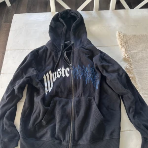 Svart hoodie från Wasted  - Svart hoodie från Wasted med stor vit och blå logga på bröstet. Tröjan har huva med snörning, dragkedja framtill och två rymliga fickor. Materialet är mjuk bomull och polyesterblandning, perfekt för chill dagar. Långärmad och loose passform.