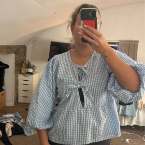 Blårutig blus med snörning från Gina Tricot - Supersöt blus från Gina Tricot i blått och vitt rutigt mönster. Blusen har puffiga trekvartsärmar och två snörningar framtill som ger en chill och trendig vibe. Perfekt för vår och sommar, lätt och luftig i bomull.