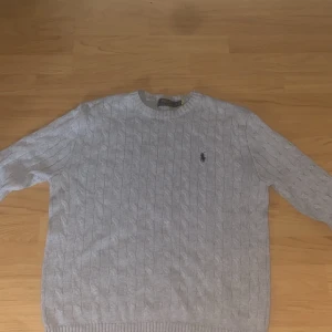 Grå kabelstickad tröja Polo Ralph Lauren - Säljer en grå kabelstickad tröja från Polo Ralph Lauren med klassisk rund hals och broderad logga på bröstet. Tröjan har långärmade och ribbade muddar, perfekt för dig som gillar stilren och tidlös design.