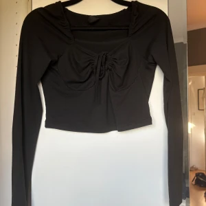 Svart croppad topp med snörning - Snygg svart croppad topp med långa ärmar och trendig snörning framtill vid bysten. Fyrkantig ringning och figurnära passform gör den perfekt till jeans eller kjol. Enkel men ändå med en cool detalj som sticker ut.