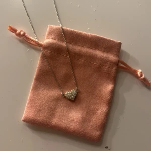 Halsband med glittrigt hjärthänge - Supersött halsband i silverfärg med ett hjärta täckt av små glittrande stenar. Kedjan är tunn och smidig, perfekt för att ge en extra touch till din outfit. Kommer med en rosa sammetsliknande påse för förvaring.