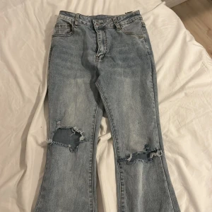Ljusblå bootcut jeans med hål - Säljer ett par ljusblå jeans med bootcut passform och snygga slitningar över knäna. Jeansen har klassisk femficksdesign, normal midja och är tillverkade i denim. Perfekta för dig som gillar en avslappnad och trendig stil. Passar S&xs