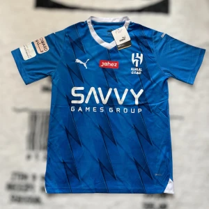 Blå Al Hilal fotbollströja Puma - Snygg blå Al Hilal fotbollströja från Puma med vit krage och tryckta loggor. Tröjan har korta ärmar, coolt grafiskt mönster och flera sponsortryck. Tillverkad i lätt och ventilerande DryCell-material som passar perfekt för träning eller match.