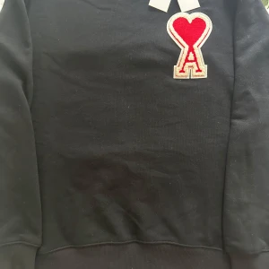 Svart sweatshirt från Ami Paris - Svart sweatshirt från Ami Paris med ikonisk röd hjärt-A patch på bröstet. Klassisk rund halsringning och ribbade muddar. Tillverkad i mjuk bomull som är skön att bära. Perfekt statement-plagg för dig som gillar stilrena detaljer.