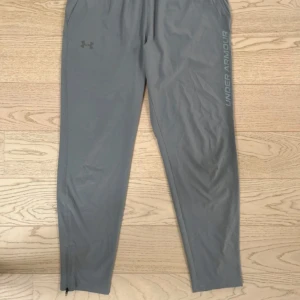 Grå träningsbyxor från Under Armour - Snygga grå träningsbyxor från Under Armour med elastisk midja och dragkedja vid benslut. Byxorna har en clean look med logga på benet och är tillverkade i ett lätt och smidigt syntetmaterial som passar perfekt för träning.