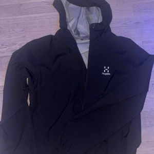 Svart Haglöfs jacka - Storlek ”L”. Skriv dm för video/prisförslag! Säljer en svart vindjacka från Haglöfs med huva och dragkedja. Jackan har ett stilrent och sportigt utseende med diskret logga på bröstet. Insidan är fodrad med mesh för extra komfort. Perfekt för blåsiga dagar och outdoor-aktiviteter.