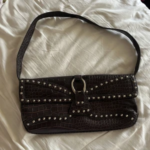 Brun axelväska med nitar och krokoprägling - Säljer en helt ny och oanvänd brun axelväska med croco-mönster och coola silverfärgade nitar på framsidan. Väskan har en avlång form, bred axelrem och snygg metallring som detalj framtill.