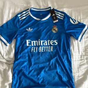 Real Madrid Tredje Ställ 25/26 (#10 Mbappé) - Snygg blå Real Madrid fotbollströja med Mbappé och nummer 10 på ryggen. Adidas logga och klubbmärke på bröstet, vita detaljer och ränder på axlarna. Rund hals och korta ärmar. Materialet är lätt och andas, perfekt för match eller träning.