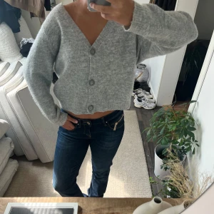 Grå croppad kofta med knappar - Säljer en ljusgrå, croppad kofta med v-ringning och tre stora knappar framtill. Tröjan är stickad och har långa ärmar, perfekt för att ge en chill och snygg vibe till din outfit. Enkel att matcha med jeans eller kjol för en trendig look. Bud på 280kr
