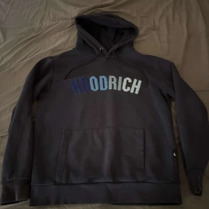 Svart hoodie från Hoodrich med brodyr - Svart hoodie från Hoodrich med broderad logga i blå och vita nyanser över bröstet. Klassisk känguruficka framtill, dragsko i huvan och långa ärmar. Perfekt för en chill och stilren streetlook.