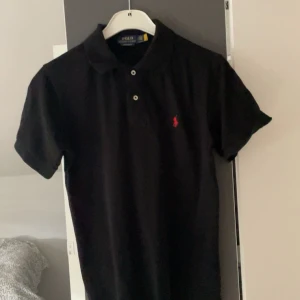 Svart pikétröja Polo Ralph Lauren - Svart pikétröja från Polo Ralph Lauren i slim fit-modell. Klassisk krage, två knappar och röd broderad logga på bröstet. Tillverkad i mjuk bomull och har korta ärmar. Perfekt för en clean och stilren look.