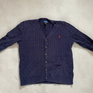 Marinblå kofta från Polo Ralph Lauren - Snygg marinblå kofta från Polo Ralph Lauren med klassiskt kabelstickat mönster och röd broderad logga på bröstet. Koftan har v-ringning, långa ärmar och knappar framtill. Perfekt för dig som gillar preppy stil och vill ha något som funkar året runt. Köpt för 1700kr mitt pris 899kr 