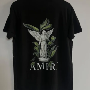 Svart Amiri t-shirt med ängeltryck - Svart t-shirt från Amiri med stort tryck av en vit ängelstaty och gröna blad på ryggen samt Amiri-logga. Framsidan har Amiri-tryck med färgade droppar. Klassisk rund hals och korta ärmar. Materialet är mjuk bomull med normal passform.