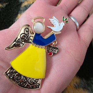 Färgglad ängel brosch med fredsduva - Charmig brosch i form av en ängel med en fredsduva. Ängeln har en gul kjol, blå topp och detaljer i guld. Den håller en skylt med texten 'Peace'. Perfekt som en symbol för fred och kärlek.