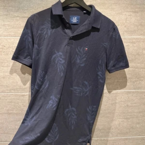 Mörkblå pikétröja från Louis Philippe - Snygg mörkblå pikétröja från Louis Philippe med diskret bladmönster. Tröjan har korta ärmar och en klassisk krage med knappar. Perfekt för en stilren och avslappnad look.