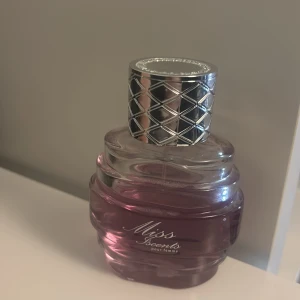 Miss Secrets pour femme parfym - Elegant parfymflaska med en stilren design. Flaskan är i glas med en lila nyans och har ett silverfärgat lock med rutmönster. Perfekt för den som vill ha en sofistikerad doftupplevelse , blommig doft