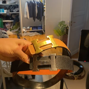Två bälten med H-spänne - Säljer två stilrena bälten med H-formade spännen. Det ena bältet är i orange och det andra i svart, båda med metallspännen. Perfekta för att ge en elegant touch till din outfit. Bara att ställa frågor och pris kan diskuteras!