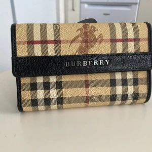 Rutig plånbok från Burberry . I ny skick  - Säljer en stilren plånbok från Burberry med det klassiska rutiga mönstret i beige, svart och rött. Plånboken har en svart läderdetalj med Burberry-loggan i silver. Perfekt för att hålla ordning på dina kort och kontanter.