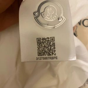 Vit pikétröja från Moncler - Säljer en stilren vit pikétröja från Moncler med klassisk krage och röd-blå randiga detaljer. Tröjan har korta ärmar och Monclers logotyp broderad på bröstet. Perfekt för en avslappnad men ändå elegant look.