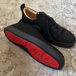 Svarta sneakers med röd sula - Snygga svarta sneakers i mocka med den ikoniska röda sulan från Christian Louboutin. Skorna har snörning och en rund tå, vilket ger en klassisk och stilren look. Perfekta för dig som vill ha en touch av lyx i vardagen.
