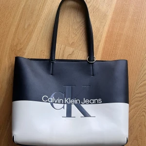 Svart och vit axelväska från Calvin Klein - Snygg axelväska från Calvin Klein. Väskan har långa handtag så att man kan ha den över axel och är stor nog för att bära med sig allt man kan behöva. Det finns gott om urymme för en laptop.