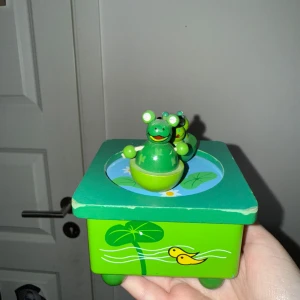 Grön speldosa med grodor - Charmig grön speldosa i trä med två dansande grodor på toppen. Basen är dekorerad med näckrosor och små fiskar . Om man vrider på den underifrån spelar den både en låt och grodorna dansar (de är magnet). Köpt second hand och sen har den bara stått och samlat damm i mitt rum!