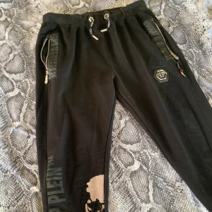 Svarta Philipp Plein byxor  - Snygga svarta mjukisbyxor från Philipp Plein med ett fett dödskalletryck med rhinestones och logotyp längs benet. Byxorna har snörning i midjan och dragkedjor vid fickorna för extra stil och funktionalitet. Lägg gärna bud! (Obs byxorna och jackan i mina inlägg är ett sett, självklart kan pris diskuteras om man vill köpa dem tsm)☺️😇