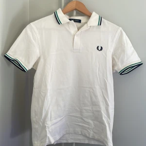 Vit pikétröja med gröna detaljer - Säljer en klassisk vit pikétröja från Fred Perry med gröna och svarta ränder på krage och ärmslut. Tröjan är perfekt för en sommar dag