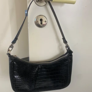 Svart axelväska med krokodilmönster - Snygg svart axelväska med krokodilmönster och dragkedja. Perfekt för att bära dina nödvändigheter med stil. Väskan har en justerbar rem och ett rymligt inre fack. Dragkedjan har tappat lite färg men det är det ända som är fel med den annars är allt fint och bra! 