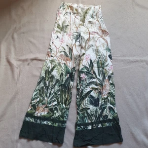 Mönstrade byxor från H&M - Supercoola byxor från H&M med tropiskt mönster i grönt och vitt. Byxorna har en utsvängd passform och är högmidjade med dragkedja i sidan. Perfekta för en somrig look med sitt djungelinspirerade tryck.