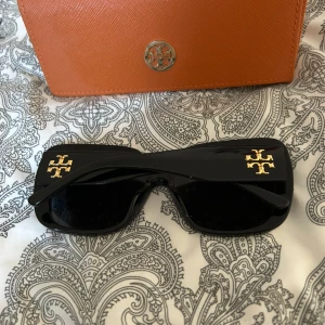 Svarta solglasögon från Tory Burch - Snygga svarta solglasögon från Tory Burch med en fyrkantig form och guldiga logodetaljer på skalmarna. Perfekta för att ge en stilren touch till din outfit.nypris-1899