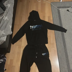 Svart tracksuit från Trapstar - Säljer en svart hoodie från Trapstar med tryck i vitt och blått på framsidan. Hoodien har långa ärmar och en klassisk huva. Perfekt för en avslappnad stil.