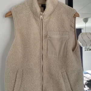  fleeceväst - Mysig beige fleeceväst från Lindex med dragkedja framtill och en praktisk bröstficka med dragkedja. Perfekt nu till våren, 